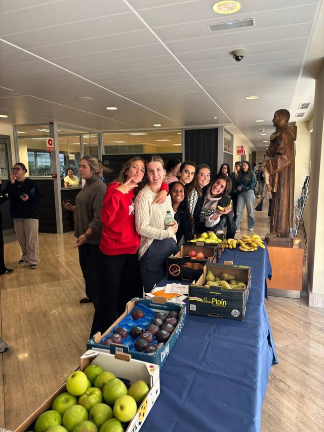 Un grupo de personas sonríe junto a una mesa llena de frutas en un entorno interior.