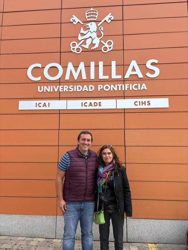 Dos personas sonrientes se encuentran frente a un edificio con el logo de la Universidad Pontificia Comillas.
