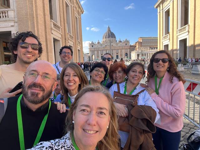 Un grupo de personas sonrientes se toma una selfie con la Basílica de San Pedro al fondo en un día soleado.