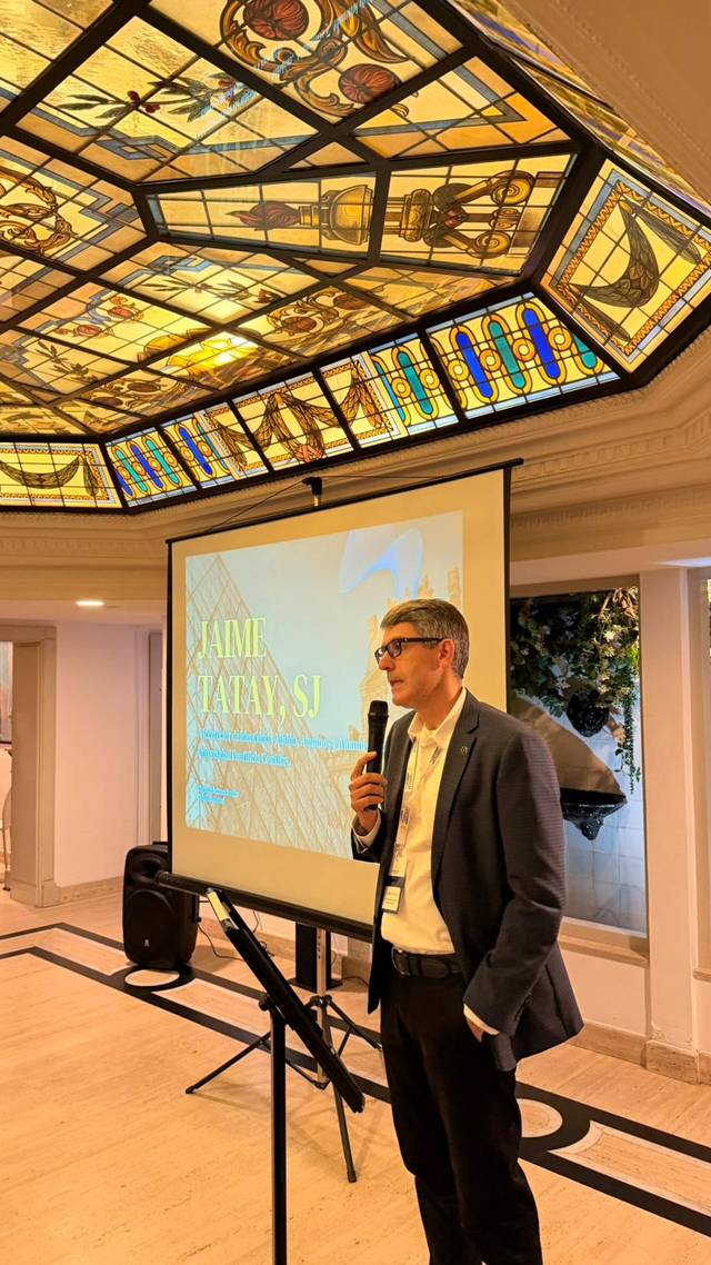 Un hombre de pie frente a una pantalla de proyección en una sala con un techo de cristal colorido.