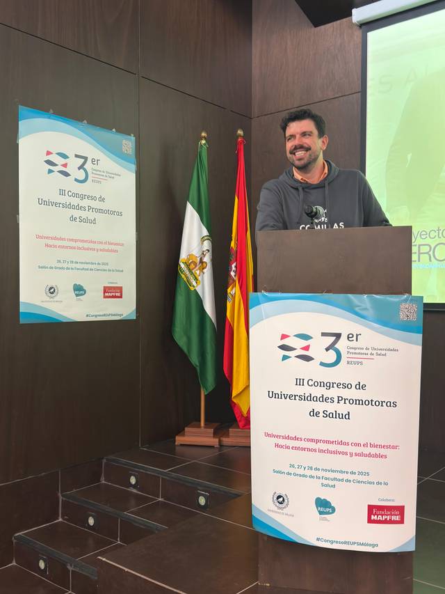 Un conferencista sonriente se encuentra de pie detrás de un podio durante un congreso sobre universidades promotoras de salud.