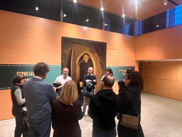 Un grupo de personas se reúne en una sala de exposiciones frente a una pintura de un personaje en un altar.