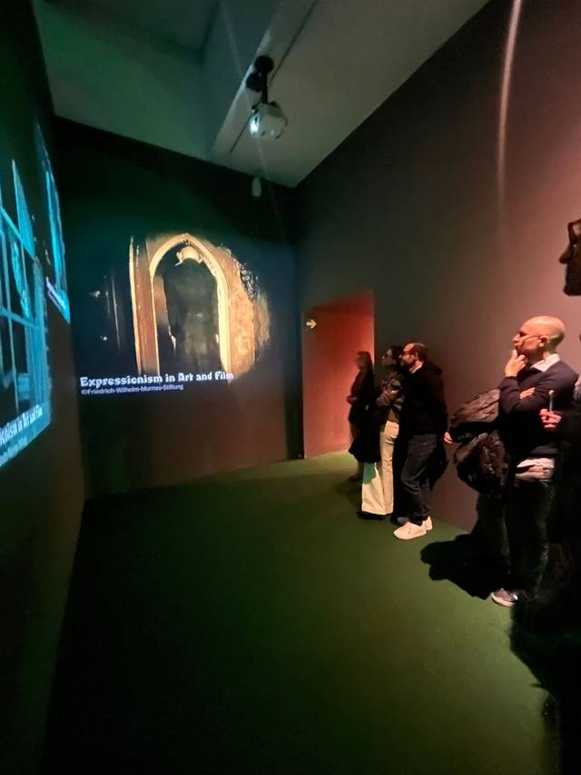 Un grupo de personas observa una proyección en una pared de una sala de exhibición de arte.