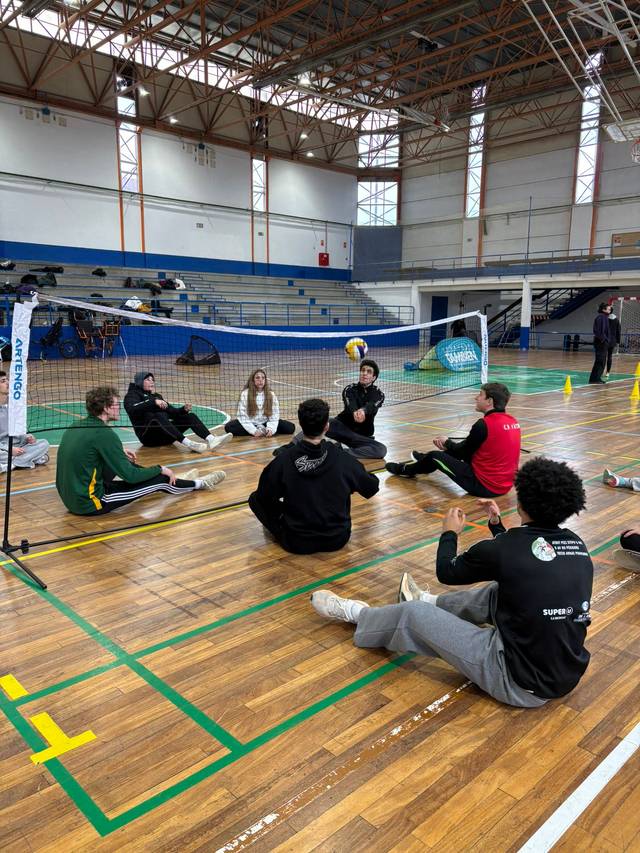 Un grupo de jóvenes se encuentra sentado en el suelo en un gimnasio, participando en una actividad relacionada con el voleibol.