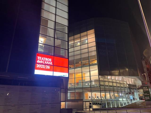 Edificio moderno de un teatro iluminado por la noche con un cartel que muestra la programación.