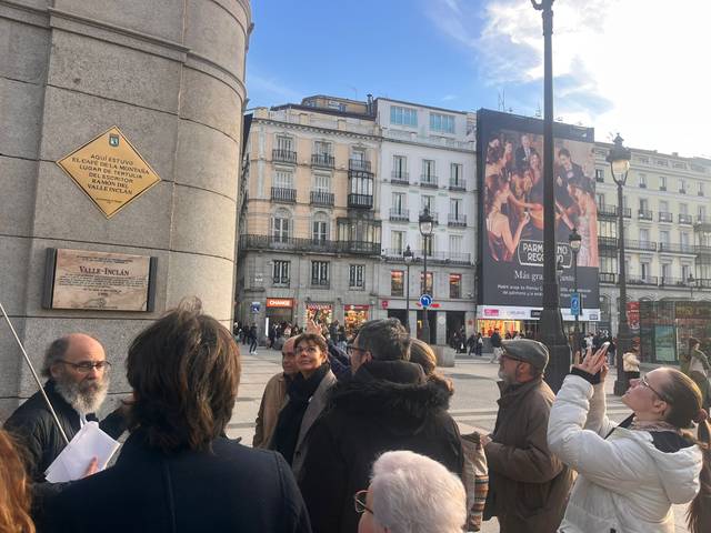 Un grupo de personas escucha atento a un guía frente a una plaza con edificios y una gran publicidad en el fondo.