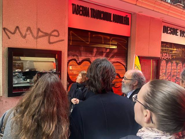 Un grupo de personas observa una vitrina mientras se encuentra frente a una taberna tradicional con graffiti en la pared.