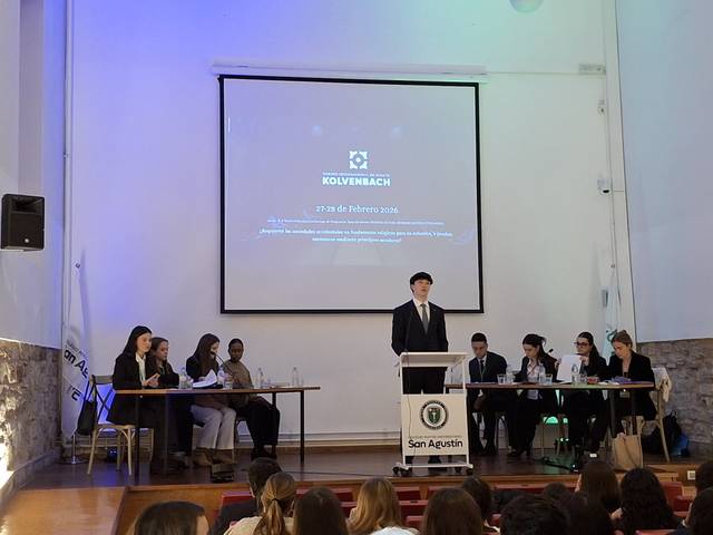 Un grupo de personas participa en un evento formal con un orador en el podio y proyectores al fondo.