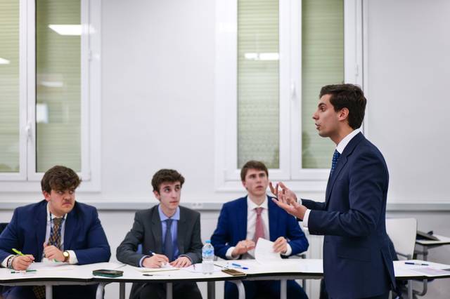 Un hombre se presenta frente a un grupo de tres personas que lo observan atentamente en un ambiente académico.