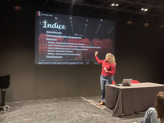 Una presentadora está explicando un proyecto en un espacio de teatro, con una proyección de diapositivas en el fondo.