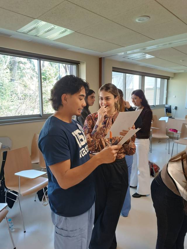 Un grupo de jóvenes discutiendo y revisando documentos en un ambiente de aula con luz natural.