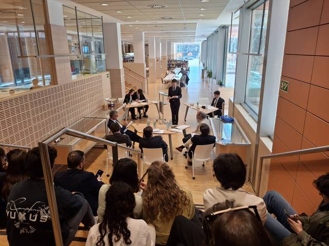 La imagen muestra a un grupo de personas sentadas en una sala en un evento formal mientras una mujer de pie parece estar presentando o moderando la discusión.