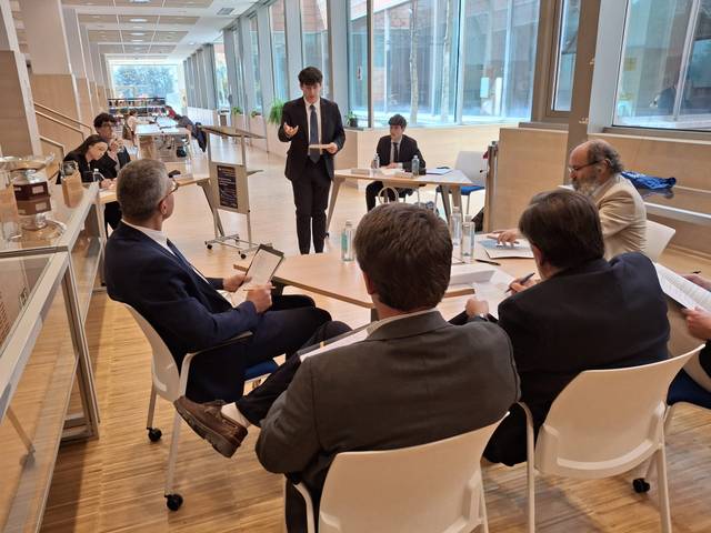 Un grupo de personas está en una sala discutiendo mientras un hombre de pie presenta ante ellos.