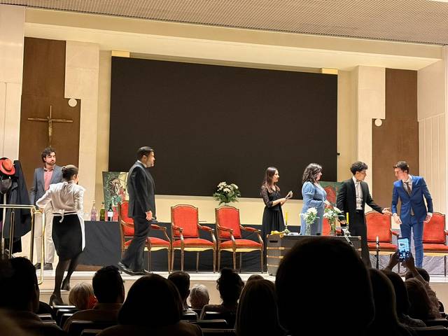Una escena teatral con varios actores interactuando en el escenario frente a un auditorio.