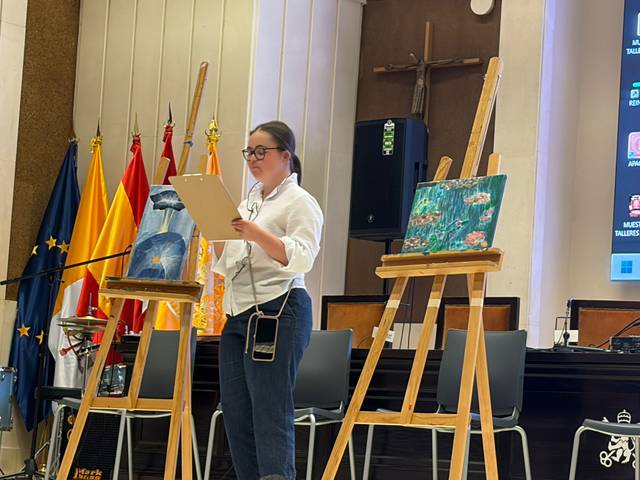 Una joven presenta su obra mientras sostiene un papel en un evento artístico.