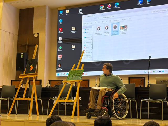 Una persona en silla de ruedas presenta su obra en un evento artístico con pantallas y obras de arte visibles.