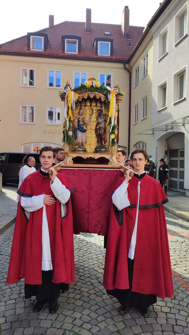 Dos jóvenes vestidos con capas rojas transportan una estructura decorativa en un evento ceremonial.