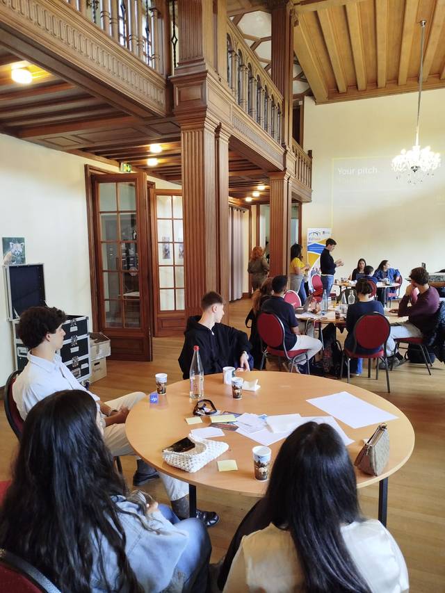 Una reunión en un salón con participantes sentados alrededor de mesas trabajando en grupo.