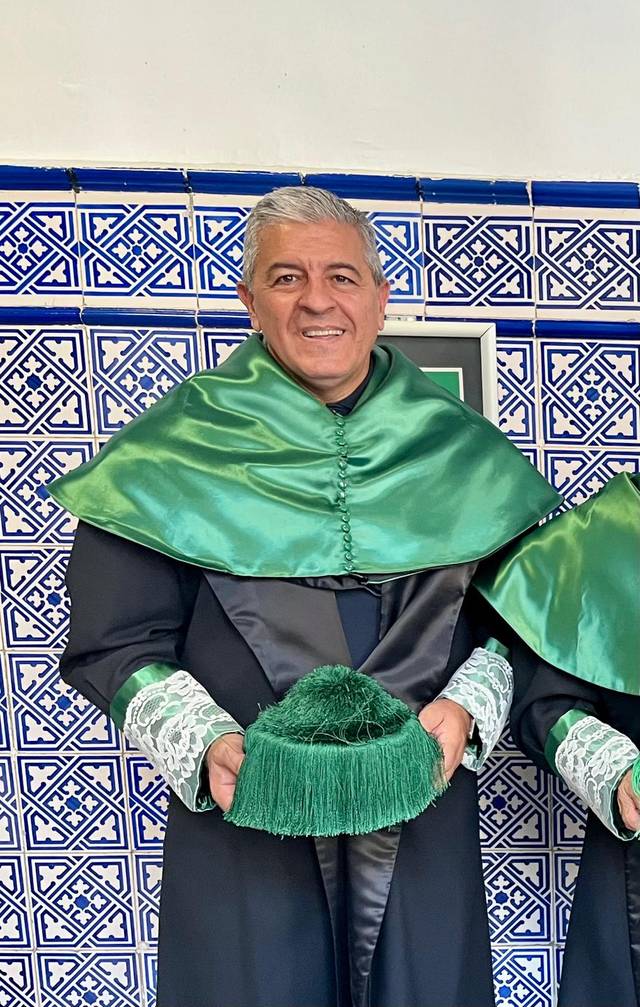 Un hombre sonriente viste una toga y capa verdes en un entorno decorado con azulejos.