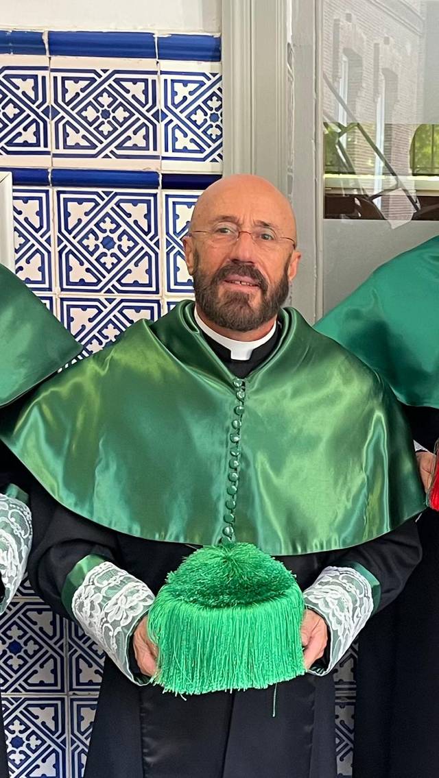 Un hombre adulto con barba y gafas, vestido con una capa verde y sosteniendo un sombrero verde, posando en un entorno decorado con azulejos.