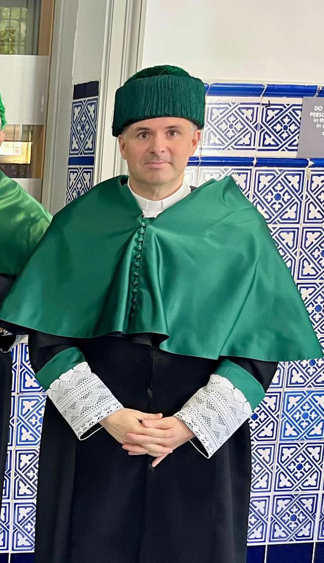Un hombre vestido con una túnica y gorro verdes, posando en un ambiente interior con azulejos.