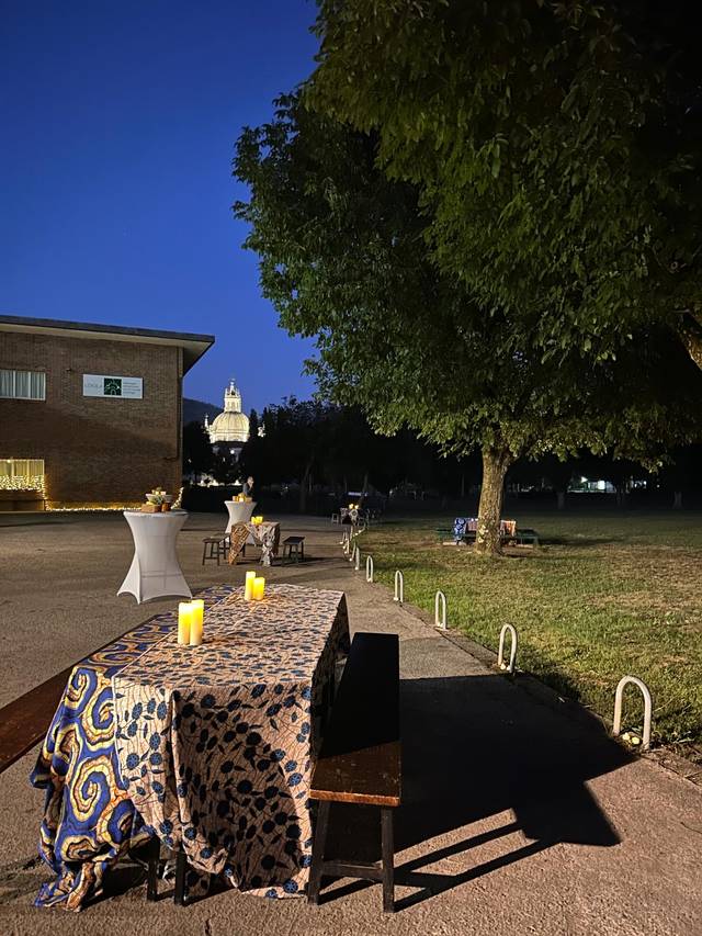 Una vista nocturna de un espacio al aire libre con mesas decoradas y un edificio iluminado al fondo.