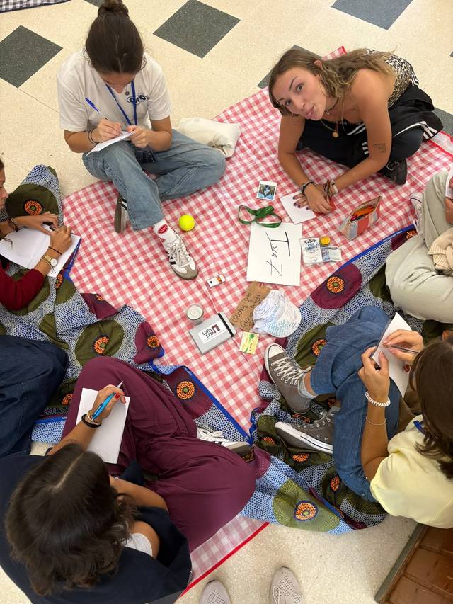 Un grupo de personas se encuentra sentadas en una manta, realizando actividades creativas y escribiendo en un ambiente relajado.