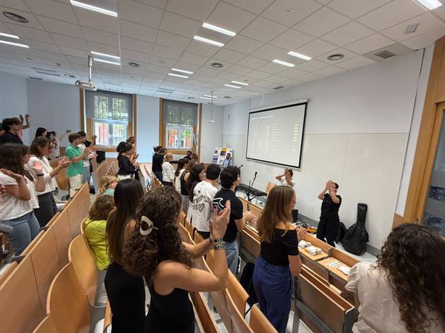 Un grupo de estudiantes se encuentra en una sala de clases, aplaudiendo a una presentadora que se encuentra en el escenario.