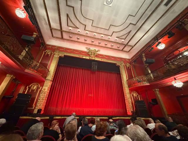Imagen del interior de un teatro con un telón rojo y una audiencia esperando el inicio de una función.