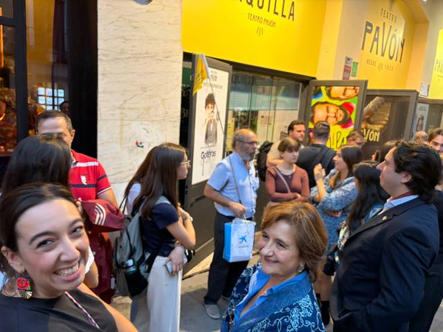 Un grupo de personas se encuentra conversando y socializando en la calle frente a un local con un cartel publicitario.