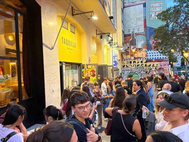 Grupo de personas esperando en una fila frente a un establecimiento en una calle con arte urbano.