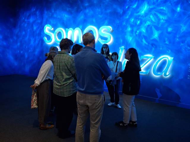 Un grupo de personas escuchando a una guía frente a una pared iluminada con la palabra 'Somos' en un ambiente azul.