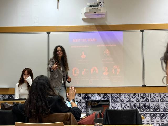 Una presentación en un aula donde una mujer de pie está hablando y haciendo una señal de aprobación mientras otra mujer la escucha sentada.