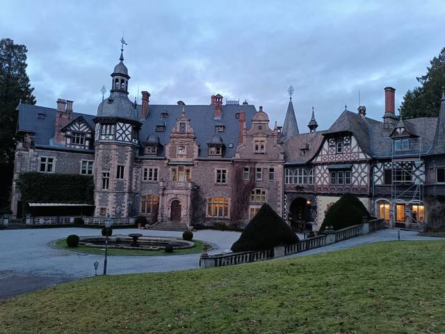 La imagen muestra un majestuoso castillo con arquitectura antigua, rodeado de jardines bien cuidados y un cielo nublado.