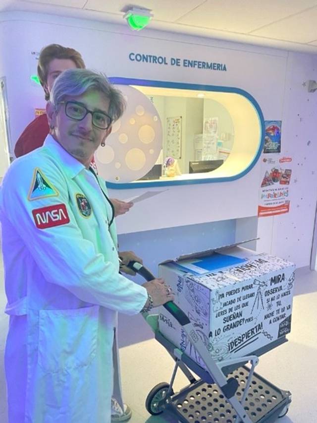 Un hombre con bata blanca y gafas empuja un carrito en un entorno que parece ser un hospital, con un fondo decorado y un letrero de control de enfermería.