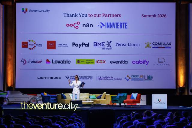 Un orador presenta en un evento mientras se muestran logotipos de diversas empresas asociadas en una gran pantalla de fondo.