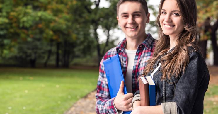 Two young students smiling and holding books in a park.