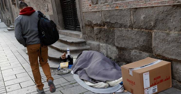 Una persona camina por la calle mientras otra está acostada con una manta en el suelo.