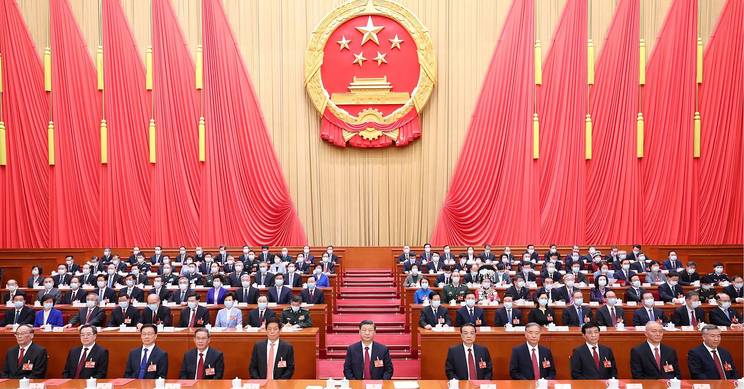 La imagen muestra una asamblea formal con una gran cantidad de personas sentadas en un auditorio decorado con cortinas rojas y el emblema de China en la parte superior.
