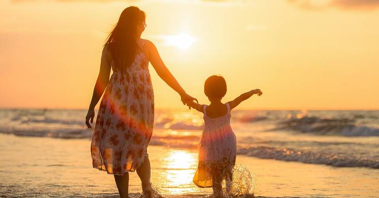 Una madre y su hija caminan de la mano por la orilla del mar al atardecer.