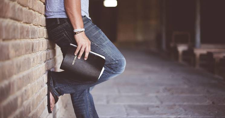 Un hombre apoyado en una pared de ladrillos con un libro en la mano.