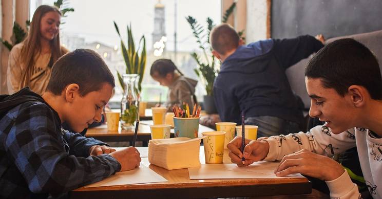 Un grupo de niños está dibujando en una mesa dentro de un café, rodeados de coloridos materiales de arte.