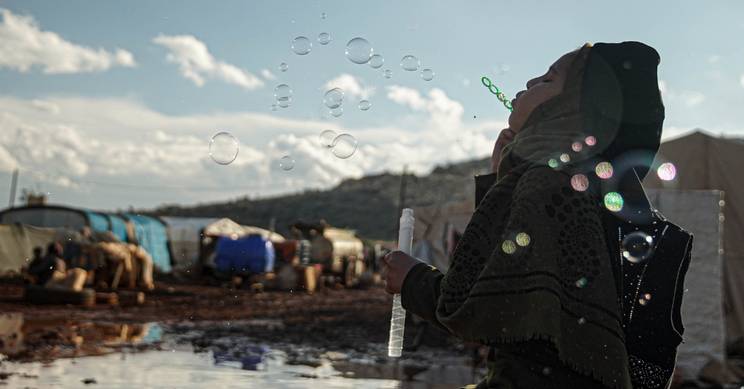 Una niña juega con burbujas en un campamento improvisado bajo un cielo despejado.