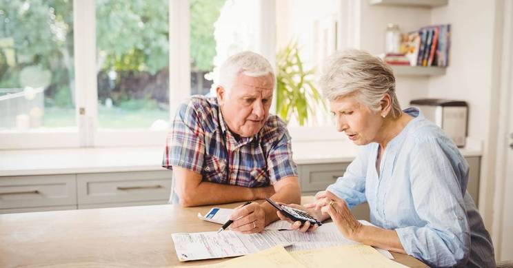 Una pareja de adultos mayores revisa documentos y un teléfono móvil en una mesa de cocina.