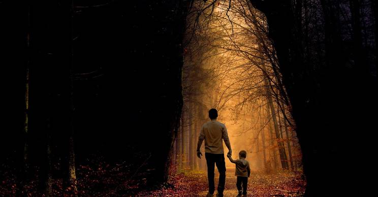 Una figura adulta y un niño caminan de la mano por un sendero iluminado entre árboles oscurecidos.