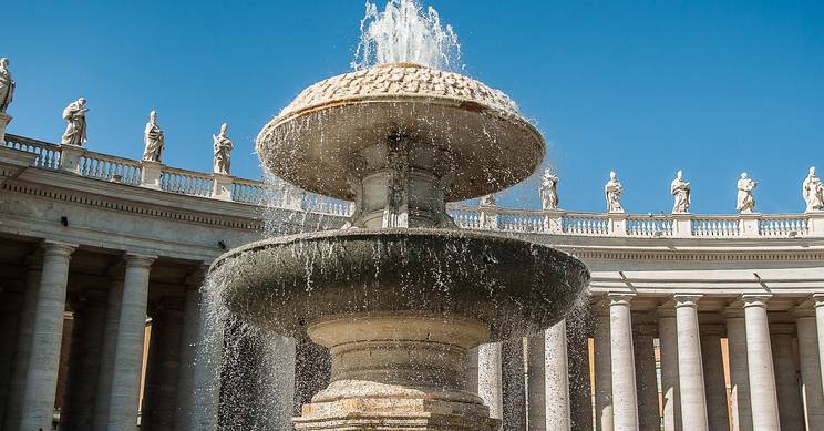 Una fuente decorativa en una plaza con columnas y estatuas al fondo bajo un cielo azul.