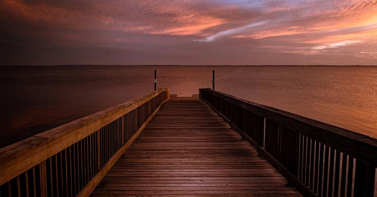 Un muelle de madera se extiende sobre un lago al atardecer, rodeado de cielos nublados y tonos rojizos.