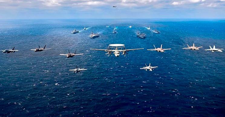 Una formación de aviones militares sobre el océano junto a varios buques de guerra.