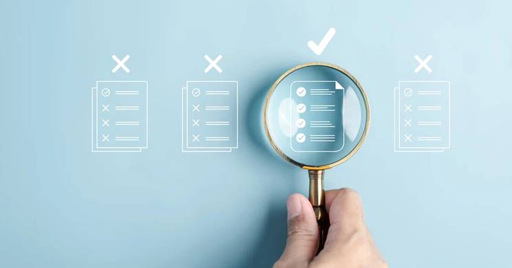 Una mano sostiene una lupa sobre documentos que muestran marcaciones y un documento con una marca de verificación.