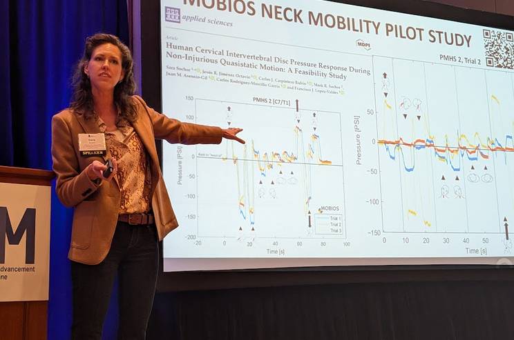 Una mujer presenta un estudio sobre movilidad cervical en una conferencia, señalando gráficos en una pantalla.